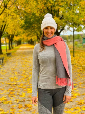 Photo for Happy Caucasian woman smiles while exploring the fall colored park on a cold November day. Cheerful female wears scarf and hat while going for a walk in park in fall - Royalty Free Image