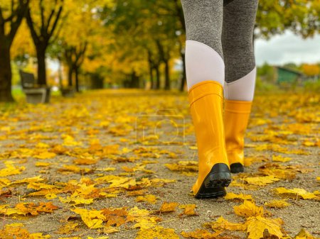 Foto de Young female explores the picturesque autumn colored park in November. Woman wearing yellow rubber boots walks along an avenue covered in leaves. - Imagen libre de derechos