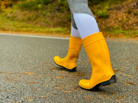 Téléchargez les photos : Woman wearing yellow rubber boots and grey yoga pants walks along a wet asphalt road. Female traveller wears boots while walking down a scenic forest route. - en image libre de droit