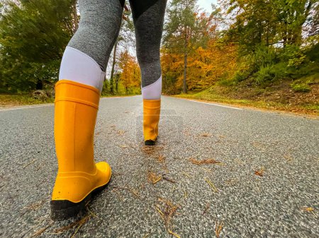 Foto de Female traveller wears rubber boots while walking down a scenic forest route. Woman wearing yellow rubber boots and grey yoga pants walks along a wet asphalt road. - Imagen libre de derechos