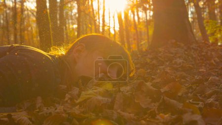 Photo for CLOSE UP, LENS FLARE: Young woman falls into pile of leaves while jogging in the autumn forest on a sunny evening. Female jogger on her morning run trips and stumbles into the dry fall colored leaves. - Royalty Free Image