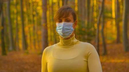 Foto de PORTRAIT, CLOSE UP, DOF: Woman wearing a surgical mask looks around while exploring the forest on a sunny autumn evening. Young Caucasian female goes for a walk in woods during covid-19 pandemic. - Imagen libre de derechos