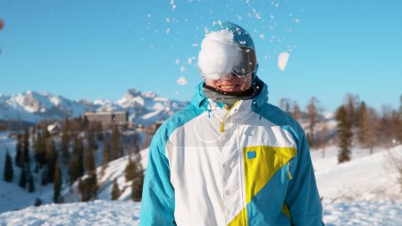 Téléchargez les photos : CLOSE UP, DOF: Stoked male snowboarder gets hit in the head by a large wet snowball. Cinematic shot of a happy young male tourist wearing goggles getting hit by a snowball during a random snowfight. - en image libre de droit