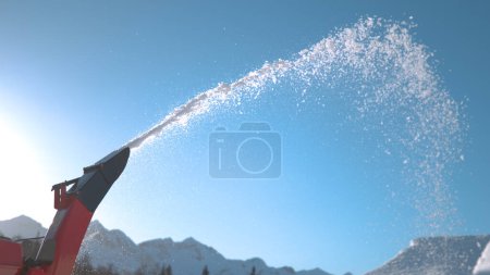 Téléchargez les photos : CLOSE UP: Detailed shot of a snowblower machine blowing snow into the sunny air. Large industrial machinery spits out small chunks of fresh snow as ski resort operators get ready for ski opening. - en image libre de droit