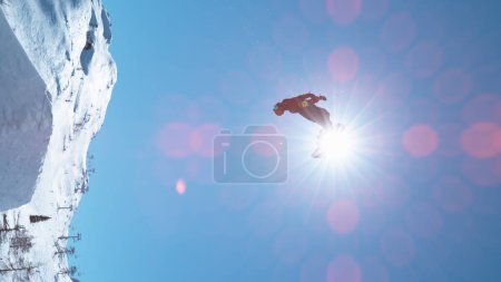 Téléchargez les photos : VERTICAL, LENS FLARE: Fearless male tourist snowboarding in fun park does a big backflip on a perfect winter day. Breathtaking shot of unrecognizable man snowboarding and doing a spectacular stunt. - en image libre de droit