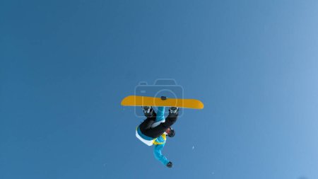 Téléchargez les photos : Male tourist snowboarding on a sunny winter day jumps off the kicker and performs a grab trick. Spectacular shot of a young snowboarder jumping high in the air and doing an extreme aerial trick. - en image libre de droit