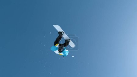 Téléchargez les photos : Fearless young male snowboarder does a breathtaking backflip after jumping off a massive kicker. Man snowboarding in the Chinese mountains performs a spectacular flip while riding in a snowboard park. - en image libre de droit