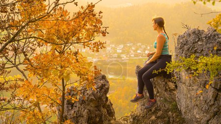 Photo for COPY SPACE: Young female hiker sits on a large rock and observes the lush green valley and emerald river below her. Woman on hiking trip stops to observe the picturesque autumn colored landscape. - Royalty Free Image