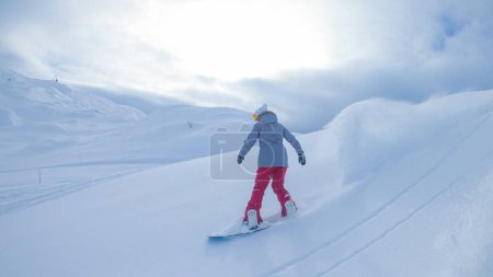 Foto de Woman snowboards in the pristine white backcountry on a beautiful December afternoon. Young female tourist snowboarding in the sunny Julian Alps of Slovenia shreds fresh powder snow. - Imagen libre de derechos