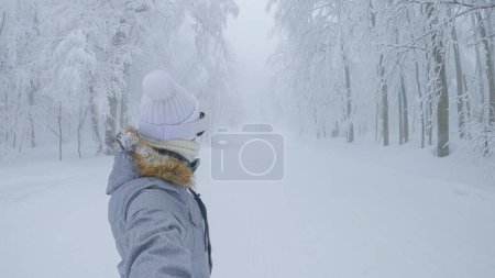 Téléchargez les photos : Athletic woman snowboards along a groomed slope leading through foggy forest. Fit female tourist rides her snowboard down an empty ski resort slope in the white wintry woods. - en image libre de droit
