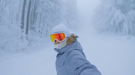 Foto de Female snowboarder with orange goggles rides along a ski resort slope leading through the misty forest. Young woman on winter vacation in the Julian Alps cruises along an empty foggy trail. - Imagen libre de derechos