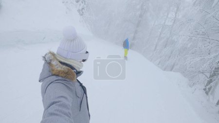 Foto de SELFIE: Los snowboarders femeninos y masculinos navegan a lo largo de una pista de esquí que corre por los bosques brumosos. Mujer joven y hombre están haciendo snowboard por una ruta forestal pintoresca en las montañas eslovenas brumosas. - Imagen libre de derechos
