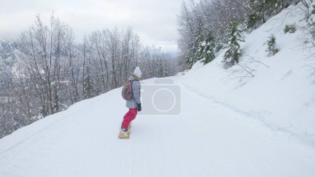 Photo for Woman snowboards along a ski resort slope leading through the forest and offering a breathtaking view of the wintry valley below. Female snowboarder rides along a flat groomed slope. - Royalty Free Image