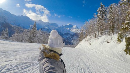 Foto de SELFIE: Unrecognizable cheerful female snowboarder cruises along a groomed slope on a sunny winter day. Young woman on active vacation in the Julian Alps snowboards along a scenic ski resort trail. - Imagen libre de derechos