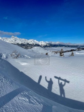 Photo for VERTICAL: Scenic shot of two excited snowboarders' shadows and wintry mountains of Slovenia. Two active tourists' shadows appear excited to be snowboarding in the Julian Alps on a sunny winter day. - Royalty Free Image
