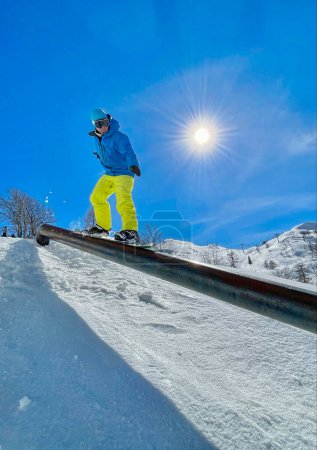 Téléchargez les photos : VERTICAL LENS FLARE: Male tourist snowboarding in Slovenia rides along a metal railing. Athletic snowboarder does an extreme railslide trick while riding in fun park of a ski resort in the Julian Alps - en image libre de droit
