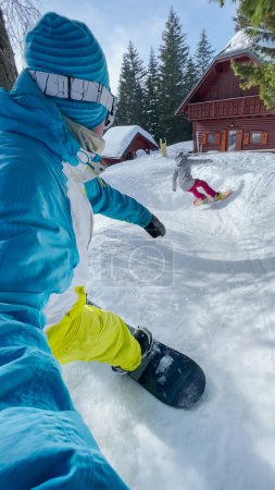 Téléchargez les photos : SELFIE, VERTICAL: Two young snowboarders ride down a trail winding between wooden cottages of an idyllic ski resort in the Julian Alps. Male and female travelers snowboard between cabin in mountains. - en image libre de droit