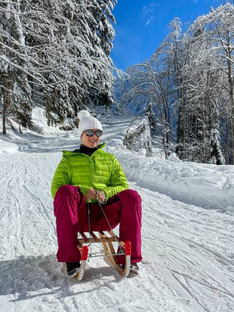 Foto de VERTICAL: Young Caucasian woman sleds down a groomed slope in the Alps on a sunny winter day. Female tourist on an active vacation in the Alps speeds down a steep slope on her vintage wooden sleds. - Imagen libre de derechos