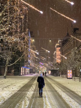 Photo for VERTICAL: Unrecognizable young woman walks down a snowy road leading through the festive city of Ljubljana on cold Christmas evening. Tourists explore the idyllic wintry streets of downtown Ljubljana. - Royalty Free Image