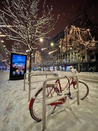 Téléchargez les photos : LJUBLJANA, SLOVENIA, DECEMBER 2020: VERTICAL: Red bike abandoned in a festive street of Ljubljana is covered in snow. Old bicycle chained to a railing is covered in fresh snow after a severe blizzard. - en image libre de droit