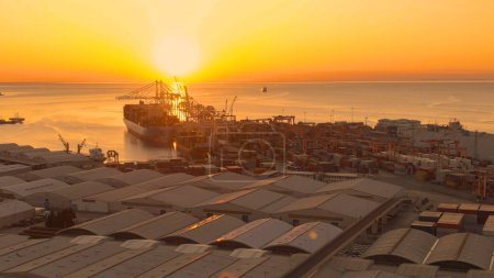 Photo for AERIAL, LENS FLARE: Picturesque shot of golden evening sunbeams illuminating the freight terminal of a large port. Breathtaking sunset shines on the cranes and cargo ship sailing toward the harbor. - Royalty Free Image