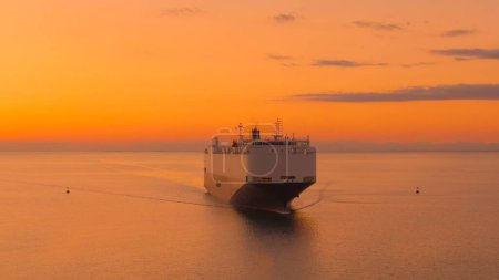 Foto de AERIAL: Golden summer evening sky spans above a freight ship transporting goods across the tranquil ocean. Scenic drone point of view of a massive cargo carrier sailing across the sea at sunset. - Imagen libre de derechos