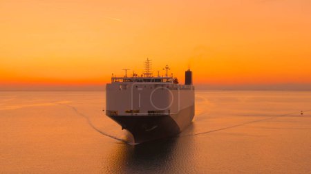 Téléchargez les photos : AERIAL: Large freight vessel is sailing across the calm ocean at picturesque burnt orange sunrise. Flying in front of an international cargo ship transporting merchandise on a sunny summer evening. - en image libre de droit