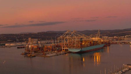 Photo for AERIAL: Flying towards a large freight ship in Koper harbor getting unloaded at dawn. Scenic shot of cranes grabbing containers off a fully loaded Maersk carrier. International port at calm sunrise. - Royalty Free Image