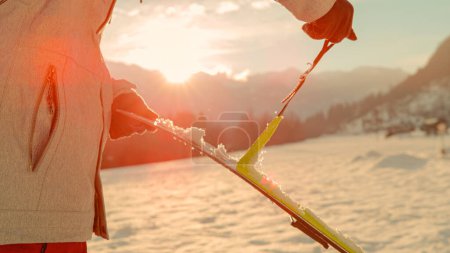 Foto de CLOSE UP, LENS FLARE, DOF: Split boarder takes the protective layer off the bottom of their new skis. Active tourist rips skins off the bottom of their split boarding gear before riding at sunrise. - Imagen libre de derechos