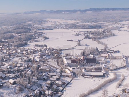 Foto de Flying high above a quiet village in the middle of wintry countryside of Slovenia. Pristine white snow covers the suburban town of Sticna on a sunny winter morning. Drone shot of snowy town. - Imagen libre de derechos