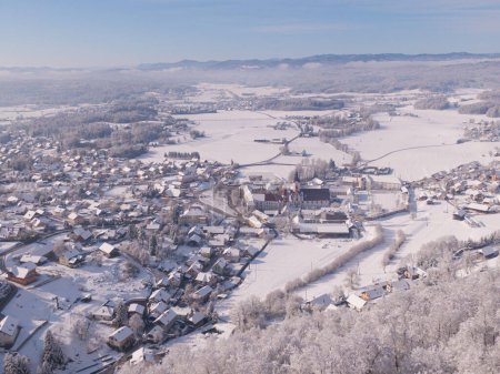 Foto de Pristine white snow covers the suburban town of Sticna on a sunny winter morning. Flying above a quiet village in the middle of idyllic wintry countryside of Slovenia. Drone shot of snowy town - Imagen libre de derechos