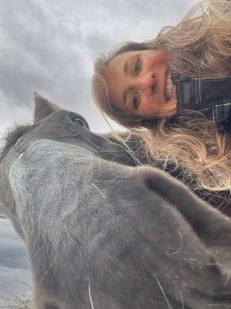 Foto de VERTICAL, SELFIE: Young female horseback rider taking a selfie with her majestic stallion on a cold winter day. Gorgeous Caucasian woman with curly hair poses next to her funny dark brown horse. - Imagen libre de derechos