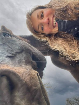 Foto de VERTICAL, SELFIE: Gorgeous Caucasian woman with curly hair poses next to her funny dark brown horse. Young female horseback rider taking a selfie with her majestic stallion on a cold winter day. - Imagen libre de derechos