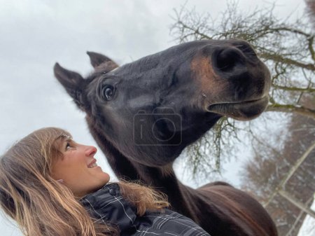 Foto de SELFIE: Young Caucasian woman lovingly looks at her beautiful dark brown horse after a long ride in the wintry countryside. Beautiful selfie shot of a female rider bonding with her majestic stallion. - Imagen libre de derechos