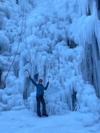 Foto de VERTICAL: Cheerful Caucasian woman ice climbing in the Slovenian mountains outstretches her arms in joy. Young female tourist is excited to start ice climbing up a breathtaking frozen waterfall. - Imagen libre de derechos