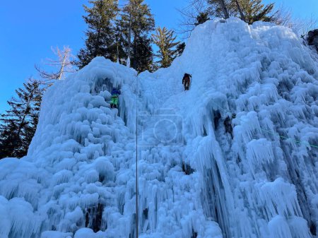 Foto de Two unrecognizable climbers ascend up a spectacular frozen waterfall in the gorgeous Slovenian mountains. Male and female alpine climbers scale a breathtaking icy wall on a beautiful sunny winter day. - Imagen libre de derechos