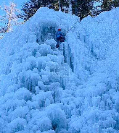 Foto de VERTICAL: Unrecognizable athletic woman ice climbs up stunning frozen waterfall. Young female tourist uses ice axes to ascend up a breathtaking light blue ice covered wall. Action shot of ice climbing - Imagen libre de derechos