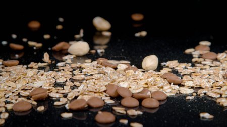 Téléchargez les photos : MACRO, DOF: Healthy muesli mixture gets scattered across an empty black kitchen countertop. A diet mix of rolled oats, blanched hazelnuts and chocolate drops falling down on an empty dining table. - en image libre de droit