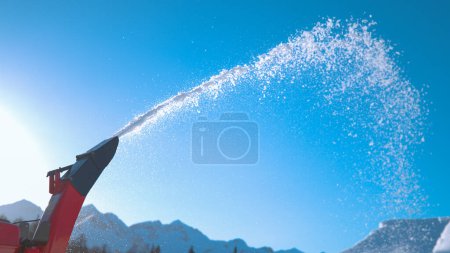 Téléchargez les photos : CLOSE UP, COPY SPACE: Industrial snow blower spews out snowflakes into the sunny sky as unknown shaper makes kickers in the snowpark of Vogel ski resort. Large industrial machine is blowing out snow. - en image libre de droit