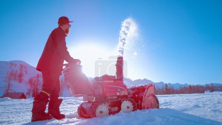 Photo for LOW ANGLE, LENS FLARE: Bright sunbeams shine on a bearded male shaper using a snow blower to create a kicker out of fresh snow. Industrial machinery operator blows snow into air on sunny winter day. - Royalty Free Image