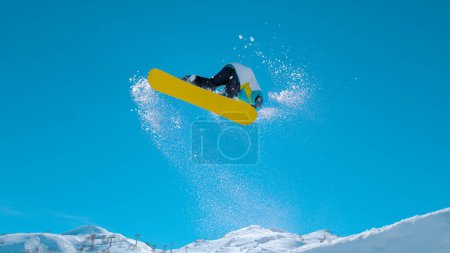 Photo for BOTTOM UP: Athletic male tourist snowboarding in the sunny Julian Alps does a spinning trick as he takes off a massive kicker. Bottom up action shot of a pro doing spectacular snowboarding stunts. - Royalty Free Image