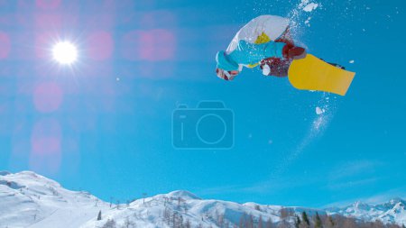 Photo for LENS FLARE: Male snowboarder jumps high in the air and does a stunning rotating stunt. Stunning action shot of a snowboarding pro doing a spinning grab trick while riding in the sunny Julian Alps - Royalty Free Image