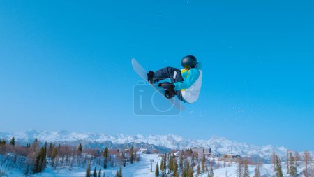 Foto de Athletic snowboarder takes off into the air and performs a spinning grab trick while enjoying a sunny winter day in sunny Vogel. Active male tourist does a snowboarding trick during winter vacation. - Imagen libre de derechos
