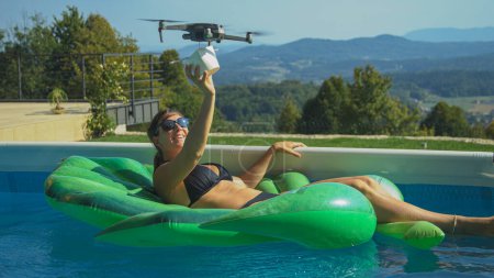 Téléchargez les photos : CLOSE UP: Drone delivers food to a young Caucasian woman relaxing in pool during the covid-19 pandemic. Female tourist on her summer vacation gets her food delivered by a contactless drone service. - en image libre de droit