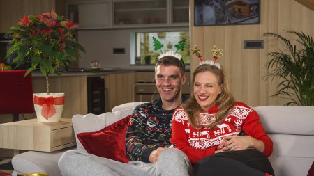 Téléchargez les photos : PORTRAIT: Cute Caucasian couple with Christmas spirit watching television on Christmas day. Beautiful young couple in love enjoying a fun Christmas movie on a couch in their decorated home - en image libre de droit