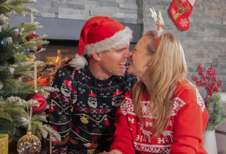 Photo for PORTRAIT: Playful young couple decorating Christmas tree with red baubles in festive holiday season. Affectionate girlfriend and boyfriend kissing and fooling around wearing Christmas sweaters at home - Royalty Free Image