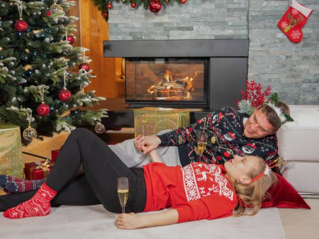 Photo for Happy young couple lying by the fireplace next to Christmas tree drinking champagne on Christmas Eve. Affectionate couple in love drinking bubbly wine while celebrating holidays at home on new year's - Royalty Free Image