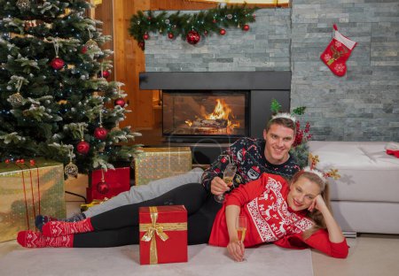 Foto de PORTRAIT: Pareja joven y feliz tumbada junto a la chimenea junto a un árbol de Navidad bebiendo champán en Nochebuena. Linda pareja bebiendo vino espumoso mientras celebra las vacaciones en casa en año nuevo - Imagen libre de derechos