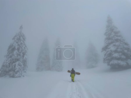Foto de Young male snowboarder walks along a narrow footpath crossing a snowy meadow in the misty Julian Alps. Man snowboarding in the Slovenian mountains treads fresh powder snow in the foggy backcountry - Imagen libre de derechos