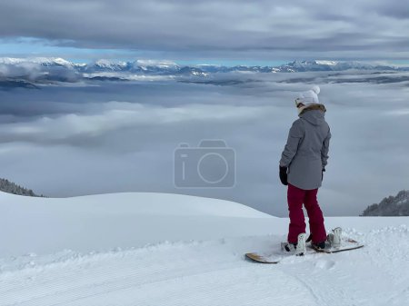 Téléchargez les photos : COPY SPACE: Athletic young woman observes the foggy valley before snowboarding in the backcountry of the picturesque Julian Alps. Active female snowboarder gazes at the breathtaking wintry mountains. - en image libre de droit